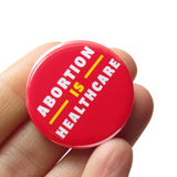 A round red pinback button that reads ABORTION IS HEALTHCARE in three lines in white & yellow text. Pin is held in a hand.