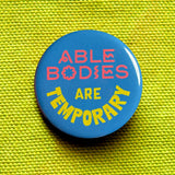 A round teal blue pinback button that reads ABLE BODIES ARE TEMPORARY in a neon red-pink & neon yellow text. Badge is pinned to yellow green fabric