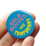 A round teal blue pinback button that reads ABLE BODIES ARE TEMPORARY in a neon red-pink & neon yellow text. Badge is held in a hand.
