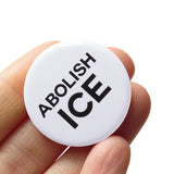 Round pinback button that reads ABOLISH ICE in black text on a white background. Button is held in a hand.