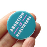 A round teal-blue pinback button that reads ABORTION IS HEALTHCARE in three lines in white & lavender text. Pin is held in hand.