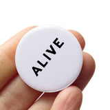 Round white 1.25" pinback button that reads ALIVE in black text. The button is held in a hand.
