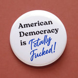 A round white pinback button that reads American Democracy is Totally Fucked in black and blue text. Button is on a clay-red background.