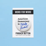 A round white pinback button that reads American Democracy is Totally Fucked in black and blue text.  Button is on a Word For Word branded backing card that reads Pinback Button, Wear Whats On Your Mind