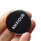 Black round 1.25" pinback button that reads ANXIOUS in a white jittery font. Badge is held in hand.