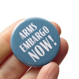 A round gray blue pinback button that reads ARMS EMBARGO NOW! in white text. Button is held in a hand.