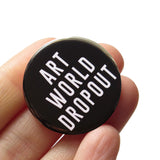 Round black pinback button that reads ART WORLD DROPOUT in white text. Button is held in a hand.