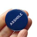 Round navy blue pinback button that reads ASSHOLE in white san serif text. Button is  held in a hand.