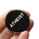 Round black pinback button that reads ATHEIST in white text . Button is held in a hand.