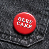 A red pinback button that reads BEEF CAKE in two lines in white serif text with a black drop shadow. Button is pinned to black denim
