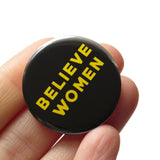Round black pinback button that reads BELIEVE WOMEN in yellow san serif text. The button is held in a hand.