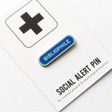 Long capsule shaped enamel pin that says BIBLIOPHILE.  Silver text and outline on a royal blue enamel background. Pin is on a Social Alert Pin backing card.