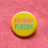 Round yellow pinback button that reads BIRTHDAY PERSON in alteranting pink & purple text, and blue and green text. with the I as a little candle. Pinned to coral denim.