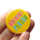 Round yellow pinback button that reads BIRTHDAY PERSON in alteranting pink & purple text, and blue and green text. with the I as a little candle. Button is held in a hand.