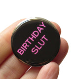 Round black pinback button that reads BIRTHDAY SLUT in pink san serif text.  Button is held in a hand.