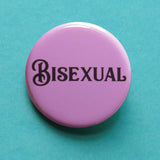 Round lavender pinback button that reads BISEXUAL in black script font. The button is on a teal background
