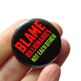A round black 1.25" pinback button that reads PBLAME BILLIONAIRES, NOT EACH OTHER in red and green text. Badge is held in a hand.