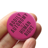 dark raspberry color pinback button that reads Bodily Autonomy Is A Human Right in dark red.Button is held in a hand.