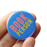 Round blue pinback button which reads BOOK PERSON  in pink and yellow text. Button is held in a hand.