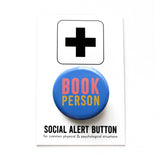 Round blue pinback button which reads BOOK PERSON  in pink and blue text. The button is on a white Social Alert Button backing card with a black plus sign at the top.
