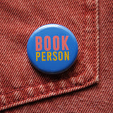 Round blue pinback button which reads BOOK PERSON  in pink and blue text. pinned to a rust-orange denim pocket.
