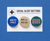 Three round pinback buttons lined up on a SOCIAL ALERT BUTTONS backing card. Reading left to right: A round blue button that reads BIBLIOPHILE in white text on two lines, A cream button that reads WELL READ in black text on two lines, and a teal button that reads VORACIOUS READER in peach & lavender on two lines.