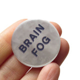 Round cloudy smokey gray 1.25" pinback button that reads BRAIN FOG in blue-gray sans serif text. Button is held in a hand.