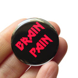 Round black 1.25" pinback button that reads BRAIN PAIN in red text. Button is held in a hand.