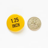 Round yellow pinback button that reads 1.25 Inch in black text. Pinback button is next to a Quarter coin for size comparison.