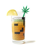 A long green acrylic laser cut drink stirrer in the shape of a pot leaf is in a crossword puzzle glass with a orange-brown beverage & a slice of lemon on the ridge.
