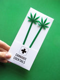 Two tall  1/8" inch thick laser cut green acrylic drink stirrers with the shape of a pot leaf cut out on top, packaged in a CANNABIS COCKTAILS, Dress Up Your Drink white & black package on a green background. The package is held at an angle with a white hand.