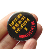 A round black 1.25" pinback button that reads Capitalism sRuns on the exploitation of workers like us in red and yellow text. Badge is held in a hand.