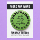 A round moss green pinback button of medium size that reads CAPITALISM in chunky black test at the top and in a black script in the middle: Sucks The Joy Out of Everything. Coins with a dollar sign fill a half moon around the edge of the button. Button is on a branded Word For Word backing card. on a lavender background.
