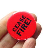 A round red pinback button that reads CEASE FIRE! in two lines in black text. Button is held in a hand.