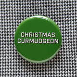 Round, dark green pinback button that says CHRISTMAS CURMUDGEON in white text with a black shadow. Button is on a tiny herringbone pattern shirt.
