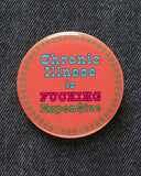 Large coral colored pinback button with a ring of green dollar signs around the edge of the button, reads CHRONIC ILLNESS IS in baby blue, FUCKING in hot pink, EXPEN$IVE in green. Button is pinned to black denim