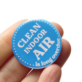 A round medium blue 1.25" pinback button that reads CLEAN INDOOR AIR, is long overdue in white text. Button is held in a hand.