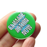 A round green pinback button that reads CLIMATE ACTION NOW in white text  with a 3D graphic effect. Button is held in a hand.