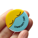 Round button with a yellow emoji face with closed eyes, wearing an aqua mask that reads COMMUNITY CARE in the shape of a smile. Button is held in hand.