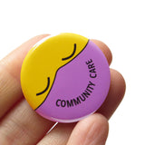 Round button with a yellow emoji face with closed eyes, wearing a lavender mask that reads COMMUNITY CARE in the shape of a smile. Button is held in a hand.