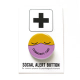 Round button with a yellow emoji face with closed eyes, wearing a lavender mask that reads COMMUNITY CARE in the shape of a smile. Button is on a Social Alert Button backing card.