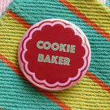 Round pink pinback button with a cookie shape that reads COOKIE BAKER in mint green text. Pinback button is on a colorful apron