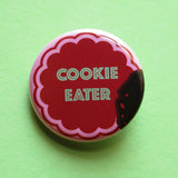 Round pink pinback button with a chocolate  cookie shape that reads COOKIE EATER in mint green text. The button looks like a bite was taken, with crumbs left behind.. Button on a mint green background.
