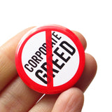 A round white button that reads CORPORATE GREED in black text with a red circle and slash to indicate being against endless greed, or END CORPORATE GREED. Button badge is held in a hand.