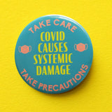 A round scrubs green pinback button that reads COVID CAUSES SYSTEMIC DAMAGE in yellow text, with peach mask icons on either side & a top curve that reds TAKE CARE, an bottom curve that reads TAKE PERCAUTIONS. Button is on a yellow background