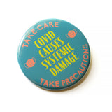 A round scrubs green pinback button that reads COVID CAUSES SYSTEMIC DAMAGE in yellow text, with peach mask icons on either side & a top curve that reds TAKE CARE, an bottom curve that reads TAKE PERCAUTIONS