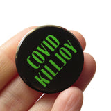 Round black pinback button with green text that reads COVID KILLJOY. Button is held in a hand.