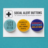 Three round pinback buttons lined up on a SOCIAL ALERT BUTTONS backing card. Reading left to right: A round aqua button reads FUCK ABLEISM, A yellow button that reads DISABILITY RIGHTS ARE HUMAN RIGHTS in black text, and a blue button that reads DISMANTLE SYSTEMIC ABLEISM in red, light blue & yellow.