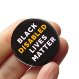 Round black 1.25" pinback button that reads BLACK DISABLED LIVES MATTER in white and yellow  text. Badge is held in a hand.