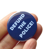 Round pinback button that says DEFUND THE POLICE. Light gray text on a navy blue background. button is held in a hand.